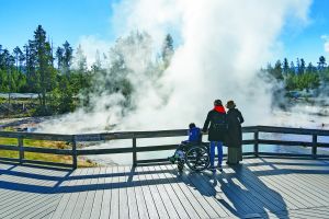 Two people with child in wheelchair on a boardwalk viewing a thermal pool.